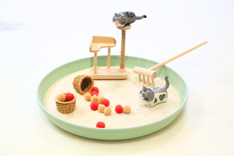 May include: A miniature zen garden with a light green tray filled with sand. It features a cat-themed design with cat figurines, a scratching post, a small rake, and decorative balls. A small basket is also included.