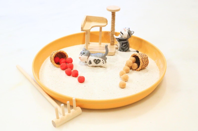 May include: A miniature zen garden scene with a yellow tray, white sand, and small cat figurines. Includes a wooden cat tree, a rake, and two baskets spilling red and tan spheres. The scene evokes a playful, miniature world.
