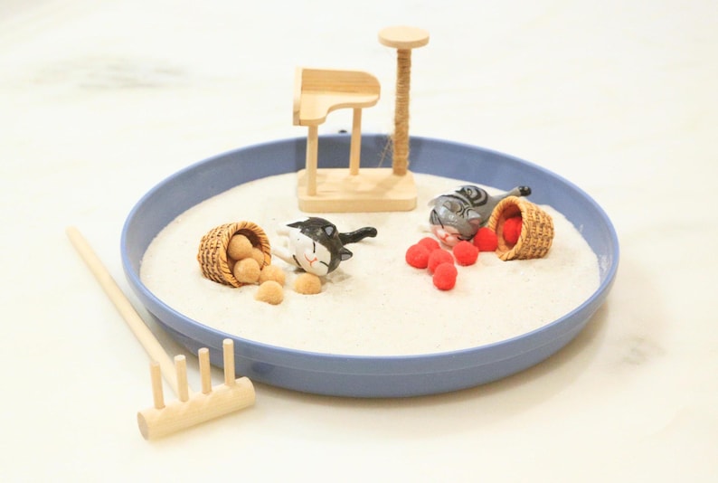 May include: A miniature zen garden in a blue tray filled with sand. It includes a wooden cat scratching post, two cat figurines, small baskets with balls and red objects, and a wooden rake. The scene is designed for relaxation and play.
