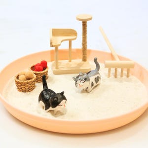 May include: A miniature cat-themed zen garden in a peach-coloured tray. The scene includes two cat figurines, small baskets with red and tan spheres, a wooden cat tree, and a rake. The sand-like material fills the tray.