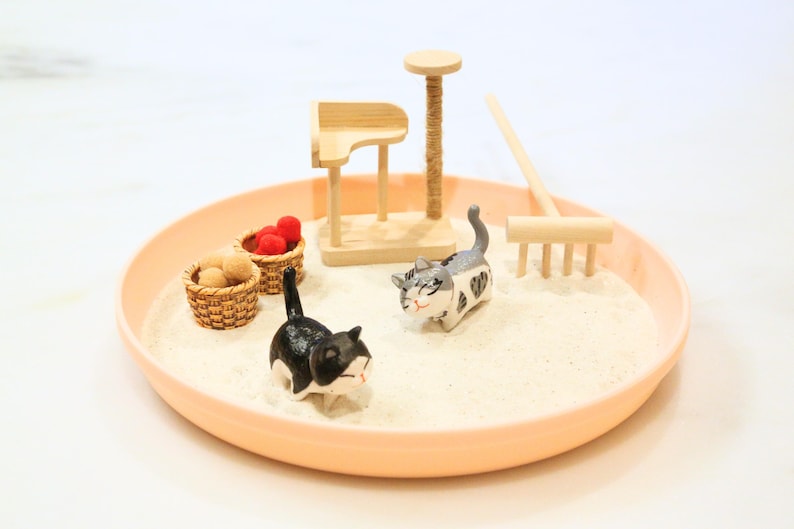 May include: A miniature cat-themed zen garden in a peach-coloured tray. The scene includes two cat figurines, small baskets with red and tan spheres, a wooden cat tree, and a rake. The sand-like material fills the tray.