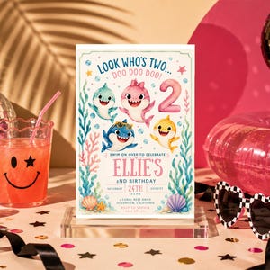Op de afbeelding: Een rechthoekige verjaardagsuitnodigingskaart met aquarelillustraties van vier kleurrijke haaien, koraalriffen en schelpen. De tekst luidt: LOOK WHO'S TWO... DOO DOO DOO! SWIM ON OVER TO CELEBRATE ELLIE'S 2ND BIRTHDAY, SATURDAY 24TH AUGUST.