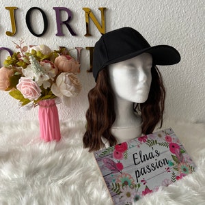 Könnte beinhalten: Eine schwarze Baseballkappe mit einem gebogenen Schirm, getragen von einer Puppe mit langen braunen Haaren. Die Puppe sitzt auf einem weißen, flauschigen Untergrund mit einem Schild, auf dem "Elna's passion" steht.