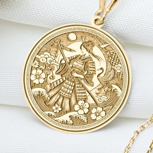 May include: Gold-colored pendant necklace featuring a detailed circular design with a samurai warrior, cherry blossoms, and waves. The pendant hangs from a delicate chain. The necklace is displayed on a white surface.