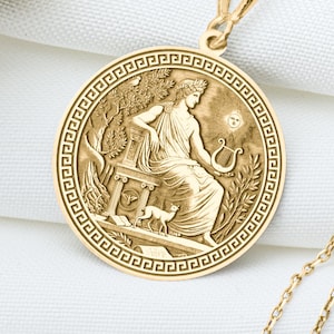 May include: Gold pendant necklace with a detailed relief of a classical figure with a lyre, set within a circular frame. The necklace is displayed on a white cloth, with dried floral accents in the background.