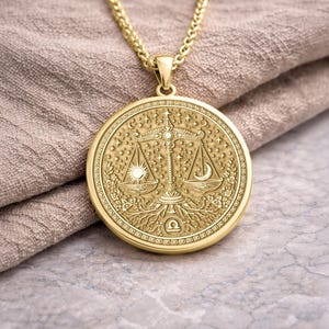 May include: Gold-colored Libra zodiac pendant necklace. The round pendant features a detailed design of the Libra scales, sun, moon, and stars. The necklace is displayed on a neutral-colored fabric.