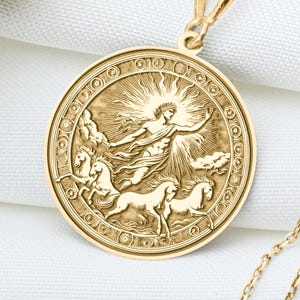 Könnte beinhalten: Eine goldfarbene Halskette mit Anhänger, die ein detailliertes Relief einer Figur mit Pferden und Sonnenstrahlen zeigt. Der Anhänger hängt an einer filigranen Kette. Die Halskette wird auf einer weißen Oberfläche mit Trockenblumen präsentiert.