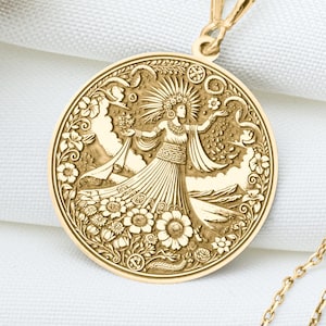 May include: Gold-colored pendant necklace featuring a detailed circular pendant with an engraved design of a woman surrounded by floral and celestial elements. The pendant hangs from a delicate chain, set against a white fabric background.