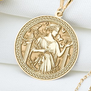 May include: A gold-colored pendant necklace featuring a circular medallion with a detailed relief of a classical figure. The figure is adorned with a laurel wreath and holds an object, with butterflies and decorative borders. The necklace is displayed on a white cloth.