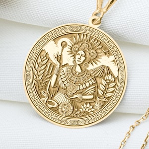 May include: A gold-colored pendant necklace featuring a detailed circular medallion. The medallion depicts a figure with intricate designs and is suspended from a delicate chain. The necklace rests on a white surface, with dried flowers in the background.
