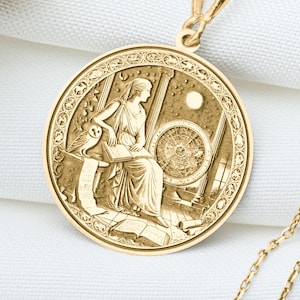 Può includere: Collana con pendente dorato con un pendente circolare inciso con una scena classica. La scena raffigura una figura seduta con un filatoio. La collana è appesa a una catena sottile. Presentata su una superficie bianca.