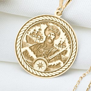 May include: A gold-colored pendant necklace featuring a detailed circular medallion with a nautical-themed relief. The medallion depicts a figure with ships and waves. The necklace chain is delicate and gold-toned, resting on a white surface.