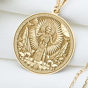 May include: A gold-colored pendant necklace featuring a detailed circular charm with a skeleton figure, sun rays, and skull motifs. The necklace is displayed on a white cloth with dried flowers in the background. The chain is delicate and the pendant is intricately designed.
