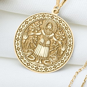May include: A gold-colored pendant necklace featuring a detailed circular medallion. The medallion depicts a figure surrounded by floral and celestial designs. The necklace chain is delicate, and the background is a white fabric.