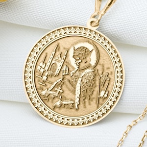 May include: A gold-colored pendant necklace featuring a detailed circular medallion. The medallion has a raised relief design depicting a religious figure and architectural elements. The necklace chain is delicate and gold-toned, resting on a white fabric surface.