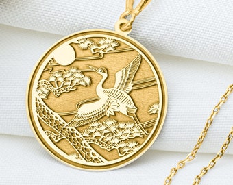 Könnte beinhalten: Goldfarbene Halskette mit einem runden Anhänger, der eine gravierte Kranich-Darstellung zeigt. Der Kranich ist vor Wolken und Sonne im Flug dargestellt. Die Kette liegt auf weißem Stoff.