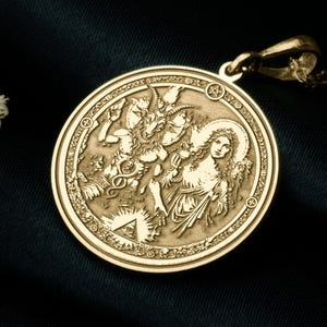 May include: Gold-coloured pendant featuring a detailed relief of a religious scene. The round pendant hangs from a delicate chain and rests on a dark, textured surface. Dried flowers and a wooden box are in the background.