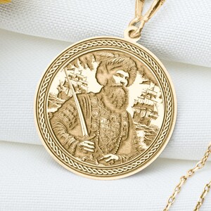 May include: A gold-colored pendant necklace featuring a detailed circular medallion. The medallion depicts a bearded figure holding a staff, with a ship in the background. The necklace chain is delicate and gold, resting on a white surface.