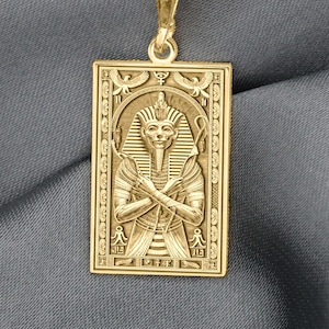 Op de afbeelding: Een goudkleurige rechthoekige hangerketting met een gegraveerde afbeelding van een Egyptische farao. De hanger hangt aan een delicate gouden ketting. De farao is afgebeeld met gekruiste armen en een hoofdtooi, tegen een gedetailleerde achtergrond.