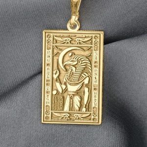 Könnte beinhalten: Goldfarbene rechteckige Anhängerhalskette mit ägyptischem Pharao-Design. Der Anhänger hängt an einer filigranen Kette. Der Pharao ist mit Kopfschmuck und Stab dargestellt, umgeben von Hieroglyphen.