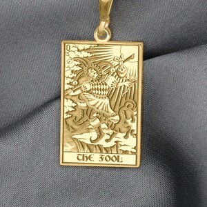May include: Gold-colored pendant necklace featuring a rectangular pendant with an engraved image of The Fool tarot card. The pendant hangs from a delicate chain. The card depicts a figure with a staff and a dog. The text "THE FOOL" is at the bottom.