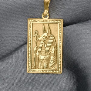 Op de afbeelding: Goudkleurige rechthoekige hangerketting met een gedetailleerd reliëf van de Egyptische god Anubis, met een staf en een ankh. De hanger hangt aan een delicate gouden ketting. De achtergrond is een grijze stof.