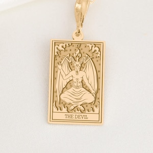 Pode incluir: Colar com pingente dourado com um pingente retangular com uma imagem gravada de Baphomet, uma divindade com chifres, de uma carta de tarô. O texto "THE DEVIL" está na parte inferior do pingente. O colar está em um fundo branco.