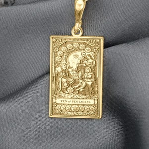 Może przedstawiać: Złoty naszyjnik z prostokątną zawieszką. Zawieszka przedstawia grawerowaną scenę z postaciami i napisem "TEN OF PENTACLES". Łańcuszek również jest złoty, a tło to szara tkanina. Biżuteria.