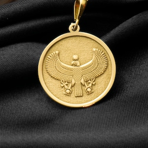 Może przedstawiać: Złoty naszyjnik z zawieszką przedstawiającą szczegółowe przedstawienie egipskiej bogini Bastet w postaci uskrzydlonego kota. Zawieszka jest okrągła i ma fakturowaną powierzchnię.