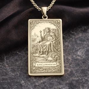Op de afbeelding: Goudkleurige hangerketting met een rechthoekig tarotkaartontwerp. De kaart toont een koning op een troon, met een scepter en bol, met de woorden "KING of PENTACLES" onderaan. De hanger hangt aan een fijne ketting.