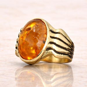 May include: A gold-toned ring featuring a large, oval amber stone. The amber has a warm, translucent orange color with visible inclusions. The ring's band has a textured design with dark accents.