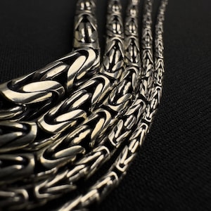 May include: Close-up of several silver-toned chain necklaces with intricate, interwoven links. The chains are arranged in a curved pattern against a dark background, highlighting the detailed craftsmanship and metallic sheen of the jewelry.