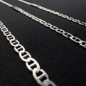 Può includere: Primo piano di tre collane a catena argentate con un design a maglie di ispirazione marina. Le catene sono esposte su una superficie nera strutturata, evidenziando i dettagli delle maglie. Catene per gioielli.