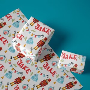 Puede incluir: Papel de regalo blanco con un patrón repetido de soldados cascanueces, hombres de jengibre, copos de nieve y bolsas de dulces. El papel tiene el nombre "Jake" impreso en letras rojas.