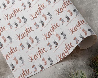 May include: White wrapping paper with a repeating pattern of colorful knitted stockings hanging from a string. The text "Have a warm & cozy Christmas" and the name "Kaylie" are repeated throughout the pattern.