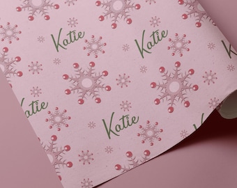 May include: Pink wrapping paper with a repeating pattern of snowflakes and the name "Katie" in green script. The snowflakes have a light pink center with red accents. The paper is partially rolled.