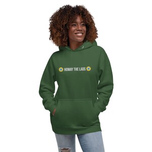 Puede incluir: Una sudadera con capucha verde con un gr&aacute;fico blanco que dice "Howay the Lads" con dos estrellas amarillas a cada lado del texto.