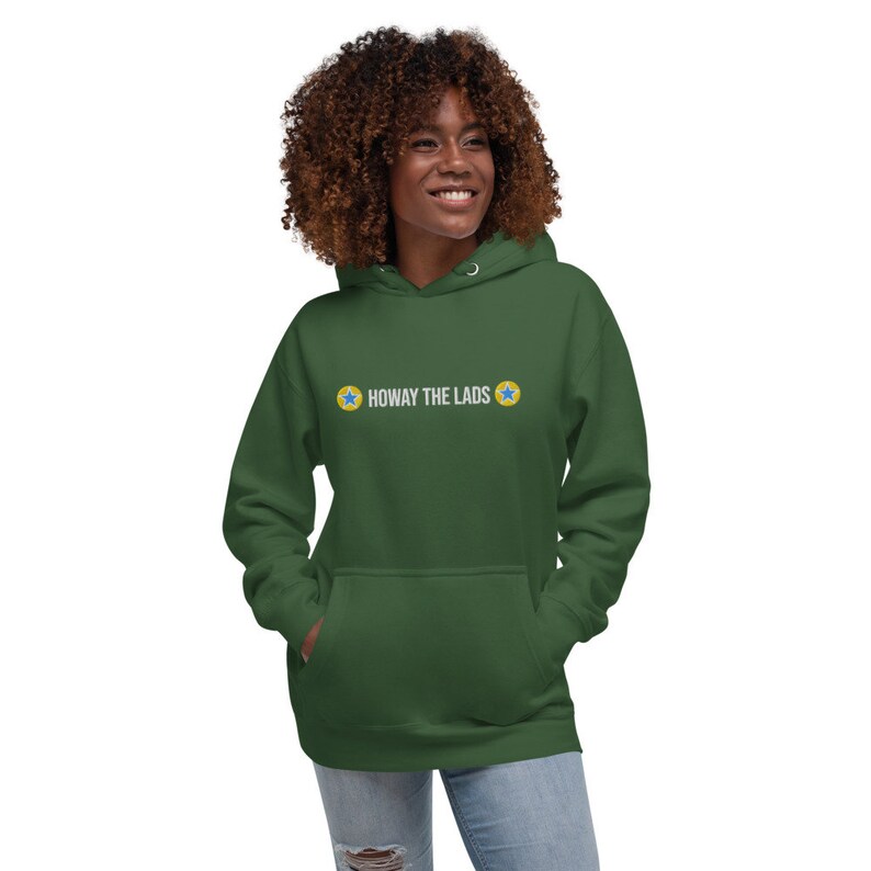 Puede incluir: Una sudadera con capucha verde con un gr&aacute;fico blanco que dice "Howay the Lads" con dos estrellas amarillas a cada lado del texto.