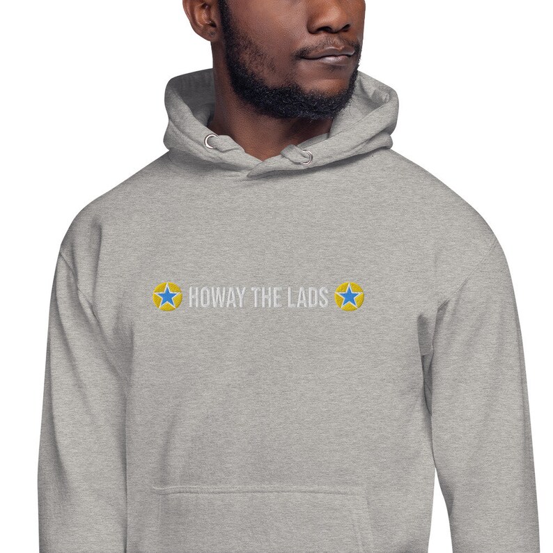 Puede incluir: Una sudadera con capucha gris con un dise&ntilde;o bordado en blanco que dice "Howay the Lads" con dos estrellas amarillas a cada lado del texto.