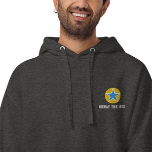 K&ouml;nnte beinhalten: Ein dunkelgrauer Kapuzenpullover mit einem blauen Stern in einem gelben Kreis und dem Text "Howay the lads" auf der Vorderseite gestickt.