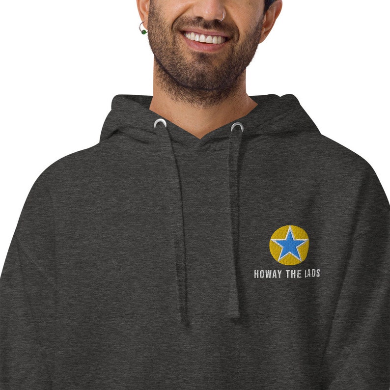 K&ouml;nnte beinhalten: Ein dunkelgrauer Kapuzenpullover mit einem blauen Stern in einem gelben Kreis und dem Text "Howay the lads" auf der Vorderseite gestickt.