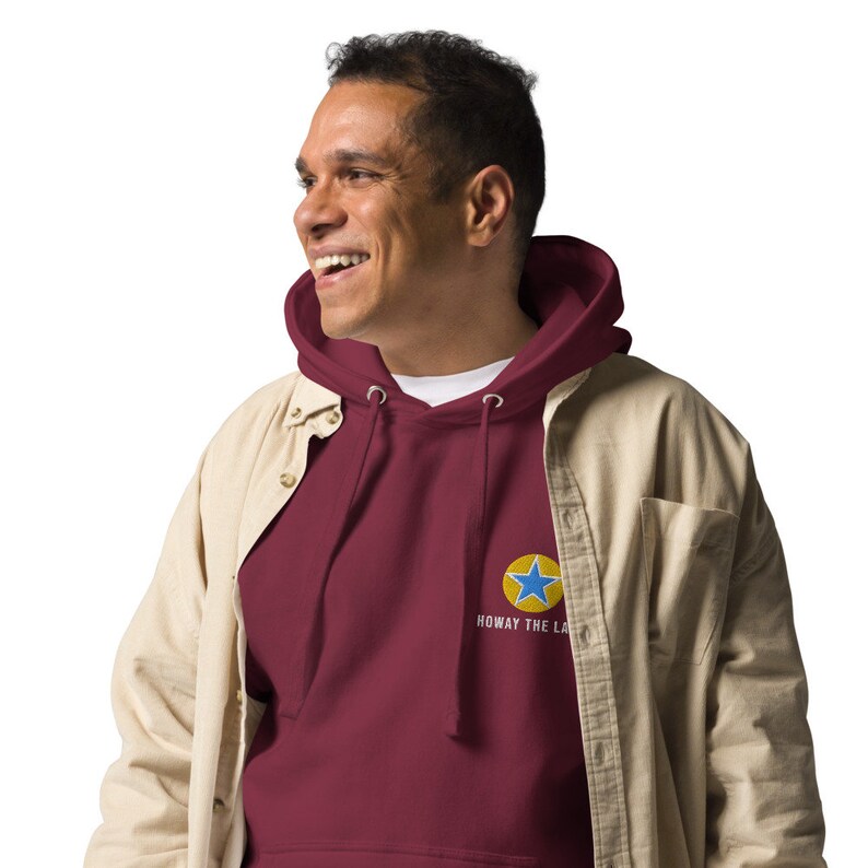 K&ouml;nnte beinhalten: Ein weinroter Hoodie mit einem gelben Kreis und einem blauen Stern. Der Text "Howay the Lads" steht unter dem Kreis.