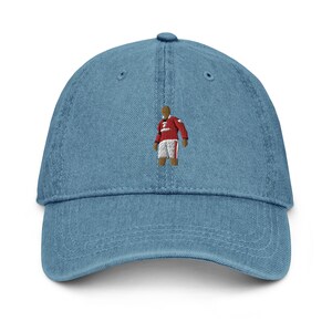 Puede incluir: Una gorra de béisbol de mezclilla azul con una imagen bordada de una persona que lleva una camiseta roja y blanca.