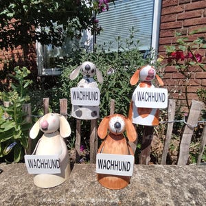 Könnte beinhalten: Vier Holzfiguren von Hunden, die jeweils ein Schild mit der Aufschrift "WACHHUND" halten. Die Hunde haben unterschiedliche Farbmuster, darunter schwarz-weiße Flecken, orange-weiße Flecken und einfarbiges Beige und Braun. Sie werden im Freien ausgestellt.