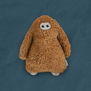 May include: A brown plush toy shaped like a monster with two black eyes. The toy has a fuzzy texture and is standing on a blue background.