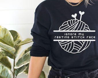 May include: A black sweatshirt with a white graphic of a ball of yarn with needles and the text "Ignore my resting stitch face".