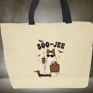May include: Cream-colored tote bag with black handles and side pockets. The bag features a Halloween-themed graphic of a ghost wearing sunglasses and a leopard print bow, holding a coffee and a purse, with a dachshund dog. The text "BOO-JEE" is above the ghost.