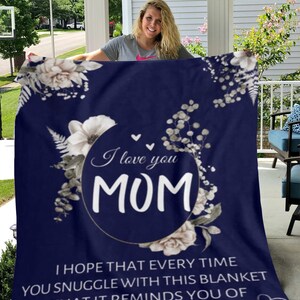 Puede incluir: Una manta de forro polar azul marino con un dise&ntilde;o floral blanco y el texto "I love you MOM" en un c&iacute;rculo. El texto debajo dice "Espero que cada vez que te acurruques con esta manta, te recuerde cu&aacute;nto te amo".