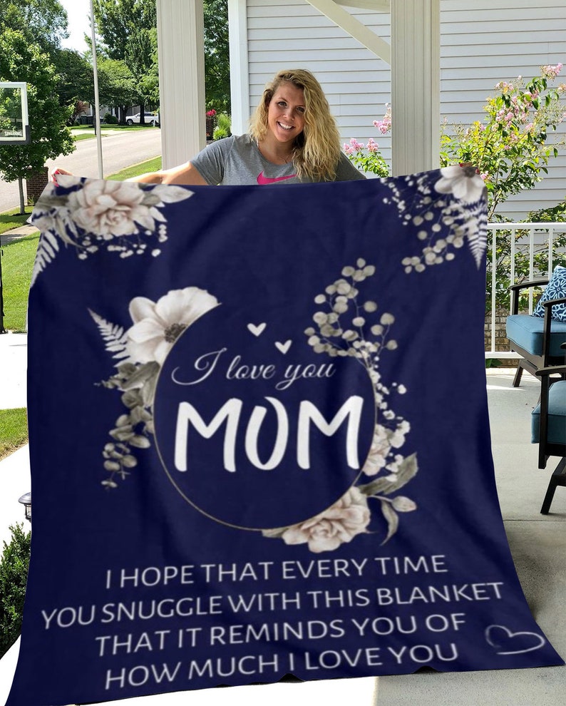 Puede incluir: Una manta de forro polar azul marino con un dise&ntilde;o floral blanco y el texto "I love you MOM" en un c&iacute;rculo. El texto debajo dice "Espero que cada vez que te acurruques con esta manta, te recuerde cu&aacute;nto te amo".