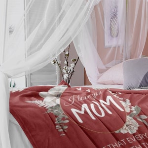 Puede incluir: Una manta de forro polar roja con un dise&ntilde;o de corona floral y el texto "I love you Mom" en un c&iacute;rculo dorado. La manta est&aacute; sobre una cama con ropa de cama blanca y un dosel.